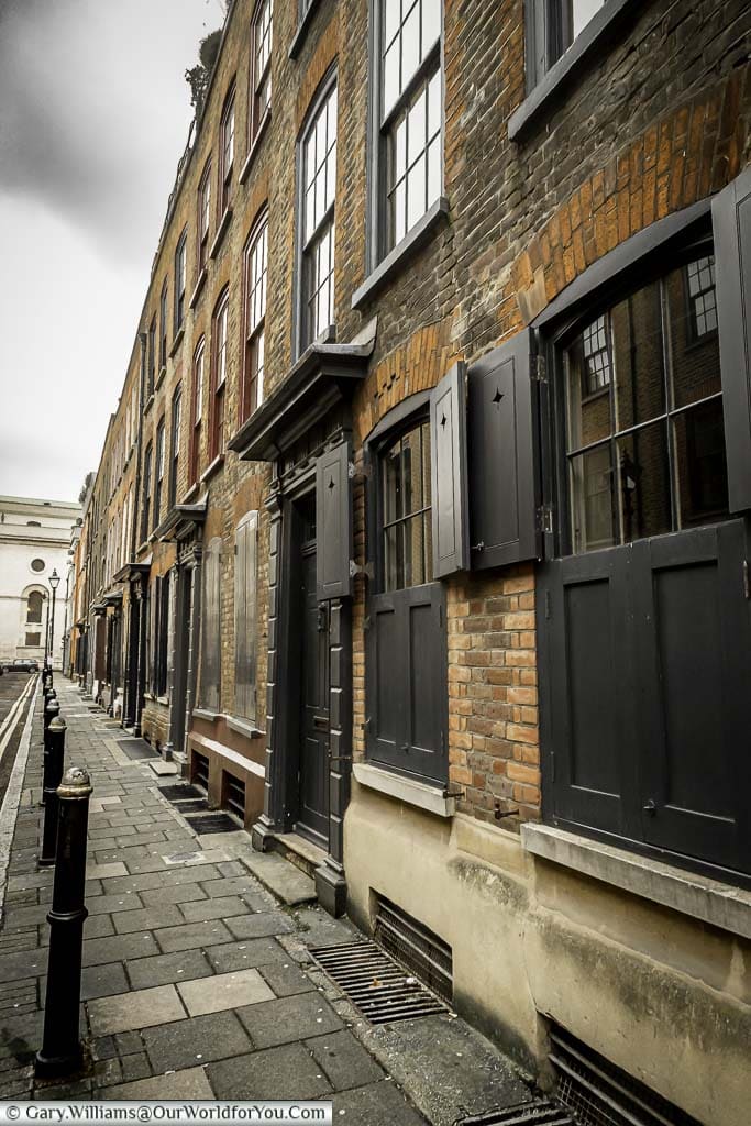 Looking along Wilkes Street, Spitalfields, a row of terraced 3 storey brick-built houses with their uniform black shuttered windows, and the pavement lined with cast iron bollards.