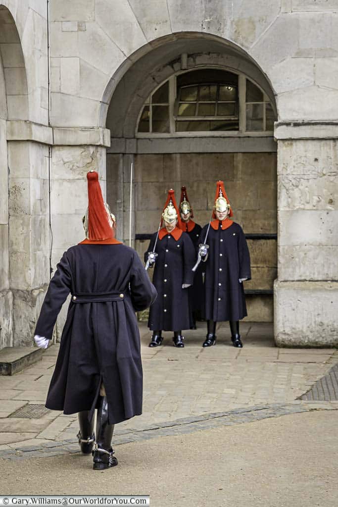 A single sentry from the Blues and Royals, in a uniform consisting of dark blue coats with vibrant red collars with red plumed helmets, marches towards three other sentries during the changing of the guard.