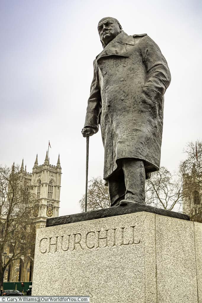 The bronze statue of Sir Winston Churchill in his later years wearing a heavy overcoat and using a walking stick, with the towers of Westminster Abbey in the background.