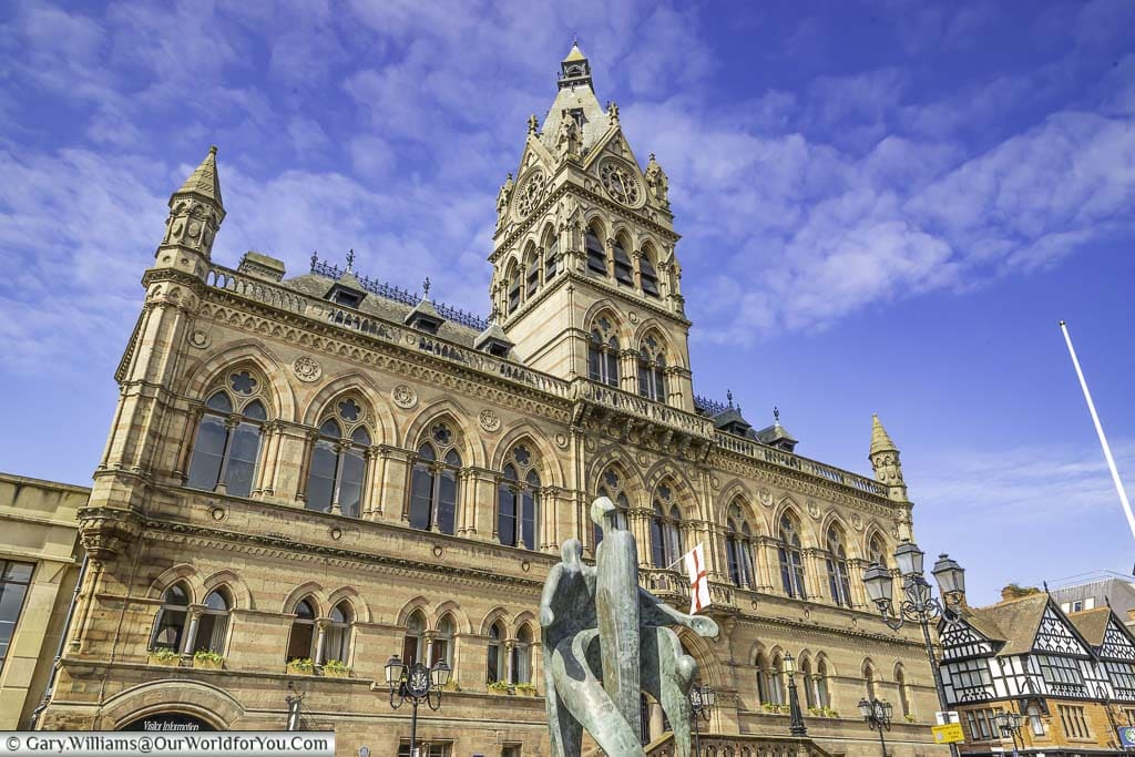 The statue 'a celebration of chester' in front of Chester's banded pink and buff sandstone townhall with a grey-green slate roof