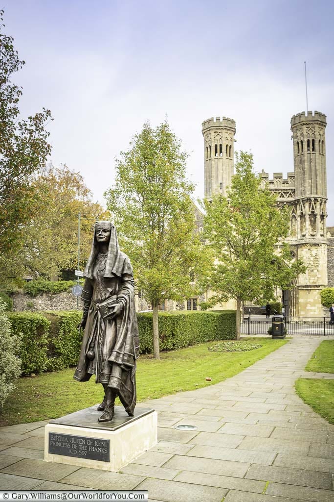 A brass statue to Queen Bertha of Kent in Lady Wootton's Green, with the gatehouse of St Augustine’s Abbey in the background.