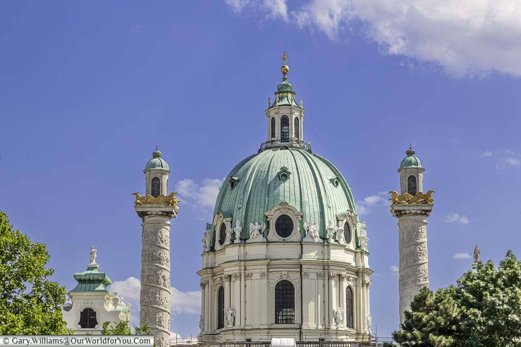 The top of Karlskirche in Vienna with its green copper-topped dome set on the white stone church. Either side are 2 decorative columns, also in white stone, topped gold trimmings and copper domes.