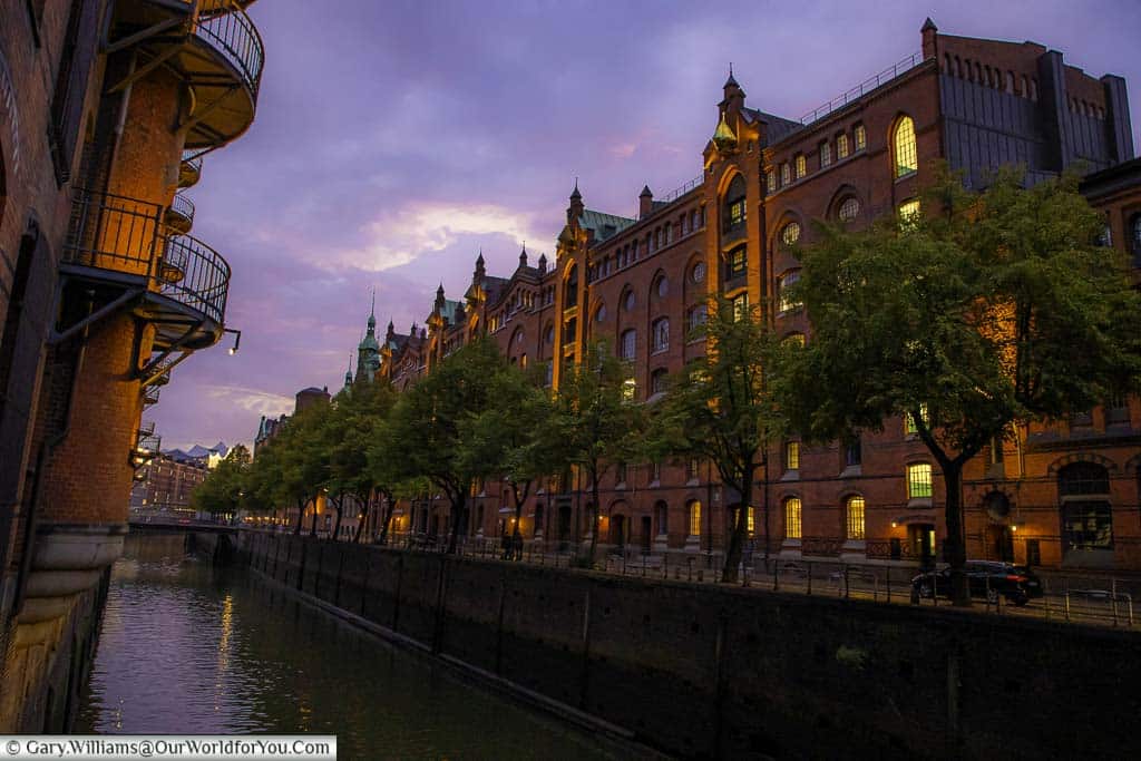 A street view of the Speicherstadt warehouse district of Hamburg at dusk with tall, imposing, illuminated red brick building under a purple sky.
