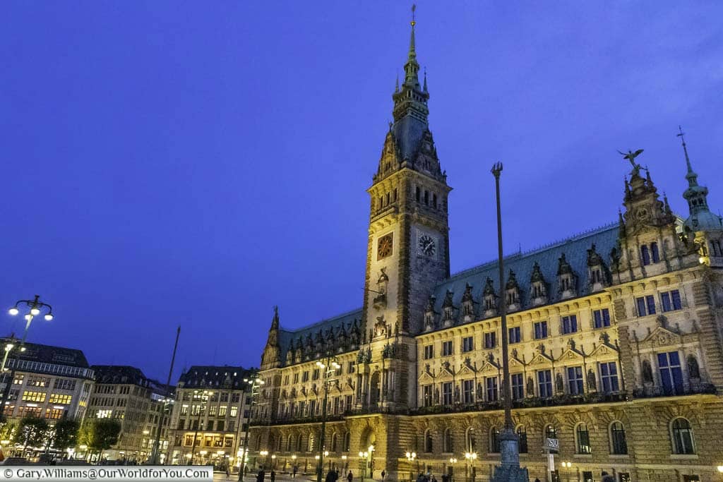 The floodlit gothic styled Rathaus of Hamburg under the blue sky of dusk, with street lights twinkling in the foreground.