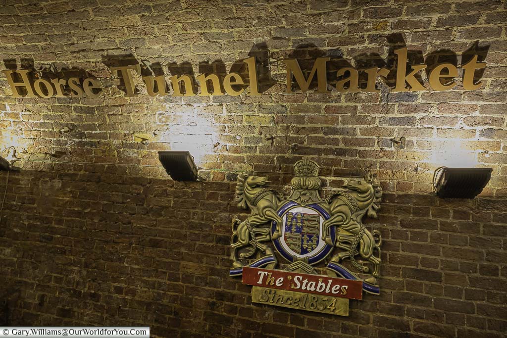 The floodlit sign for the Horse Tunnel Market within Camden Market below which is a crest between two horse figures with the label '"The Stables since 1854" below.