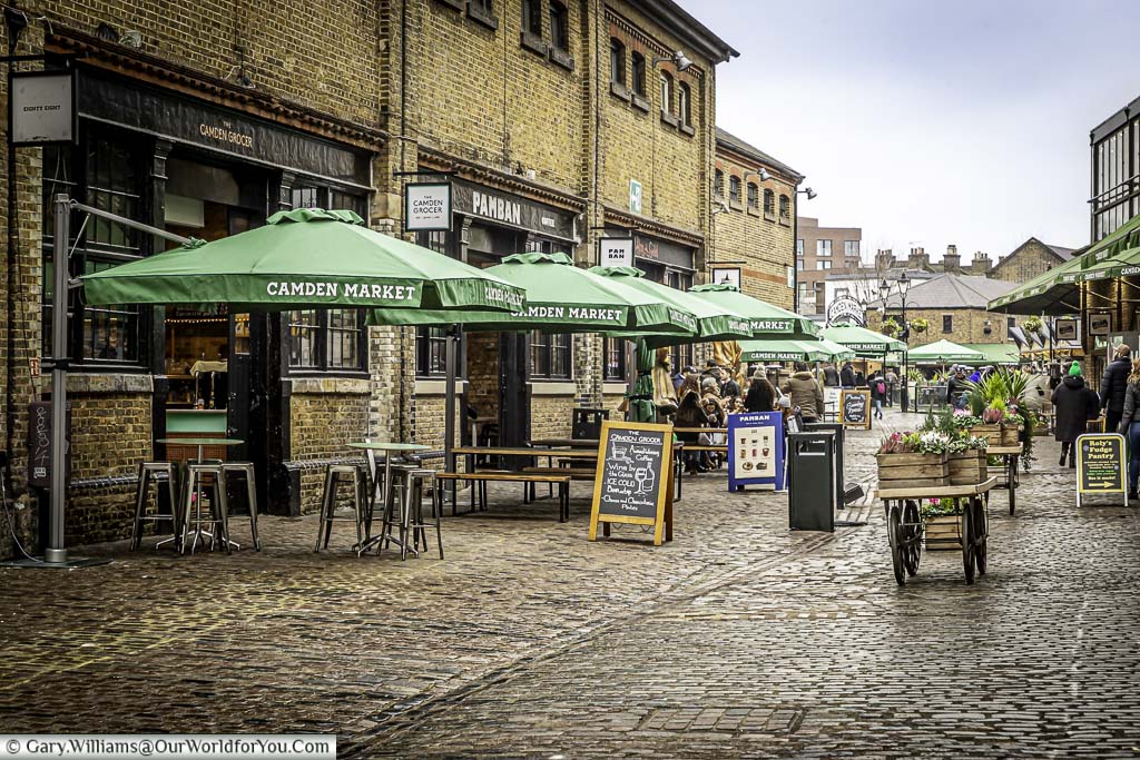 The cobbled courtyard of the North Yard of Camden Market, home to artisan food stores with parasol covered benches outside.