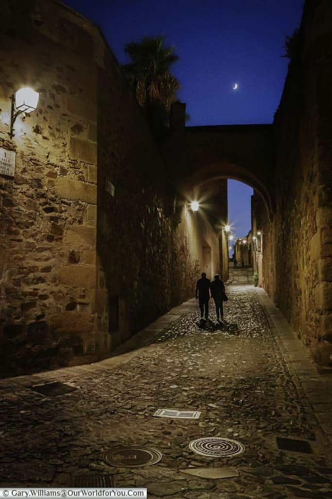 A couple walking a quiet lane in Cáceres lit by lanterns .