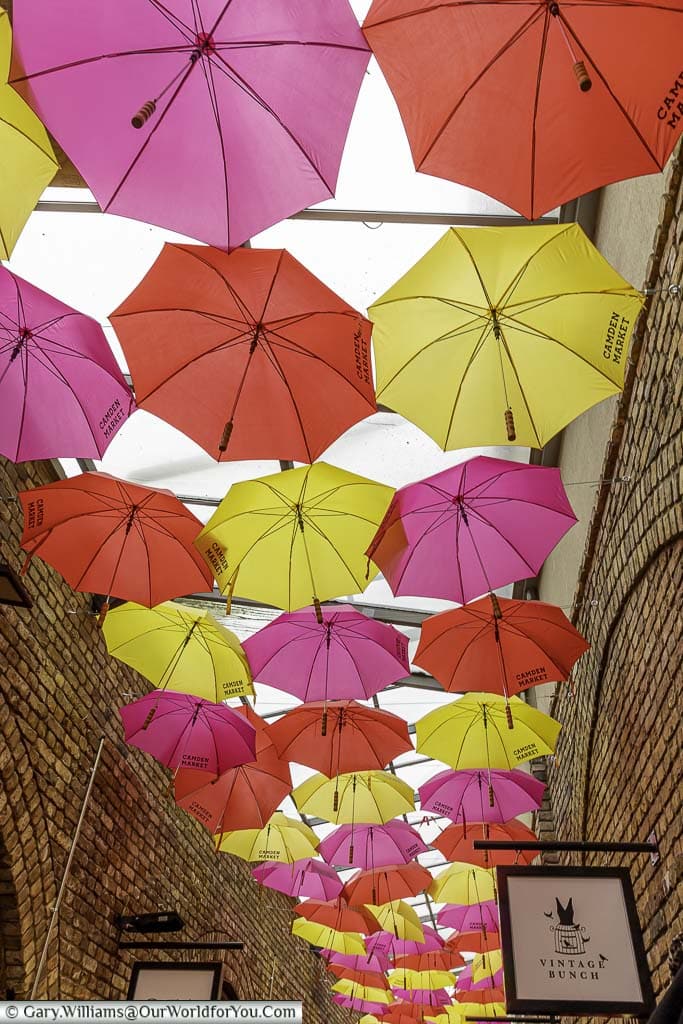 A mixture of pink, yellow & red opened umbrellas provide a canopy between the stores in one lane in Camden Market.