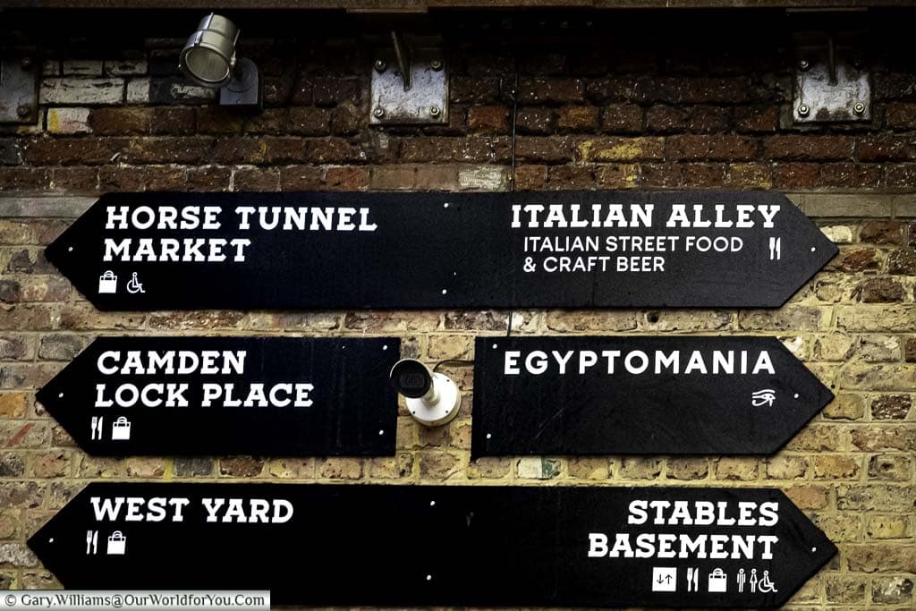 Black signs, with a white font, on a brick wall in Camden Market pointing out the key areas, including Horse Tunnel Market, Camden Lock Place, West Yard, Italian Alley, Egyptomania & the Stables Basement.