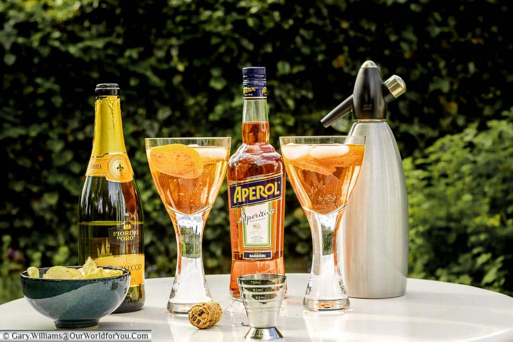 Two glasses of Aperol Spritz with a bottle of Prosecco, a bottle of Aperol, a soda syphon and a bowl of crisps in our garden