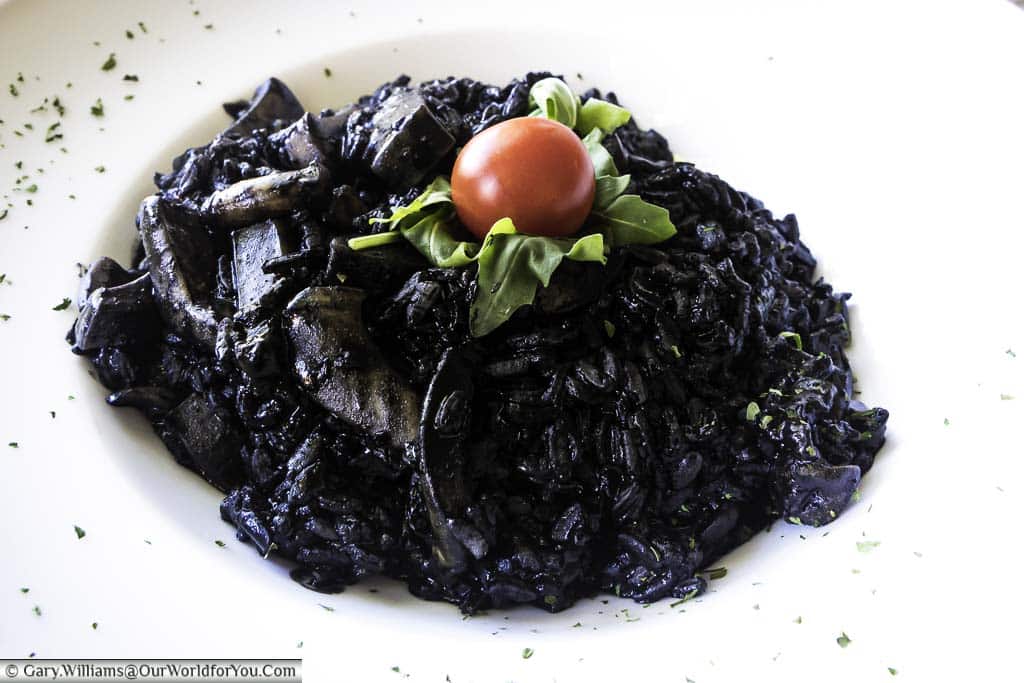 An ink-black cuttlefish risotto, topped with a green salad leaf, and a bright red tomato