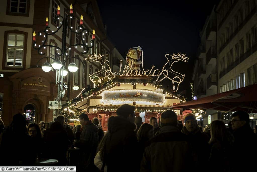 A group of people gathered around a glühwein stall on the Christmas market at night. The streel lamp has an additional advent candlelight attached, and the top of the booth has two illuminated reindeer either side of Santa and a glühwein sign.