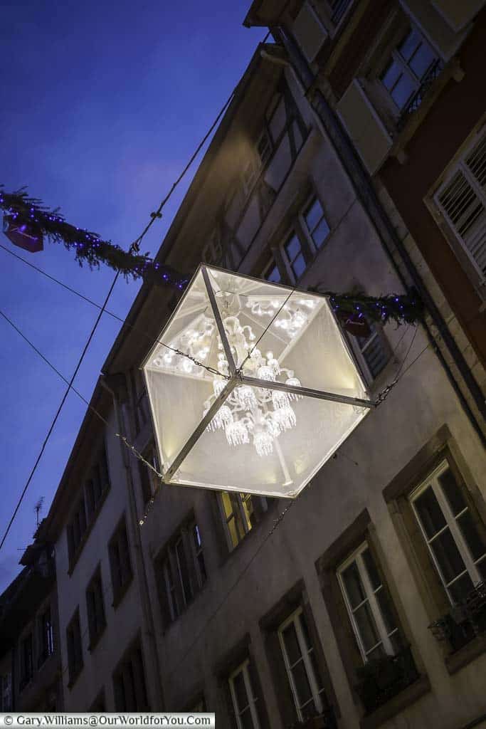 A beautiful chandelier, encased in a translucent cube suspended between two sides of the street, acting as a regular street light.