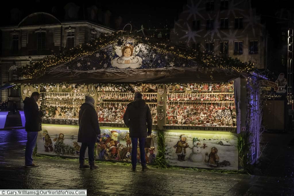 A Christmas market stall in the Place De La Cathédrale decorated with Christmas scenes of Santa and his sleigh, children with a snowman and an angel looking down at the shoppers perusing its extensive range of Christmas decorations.