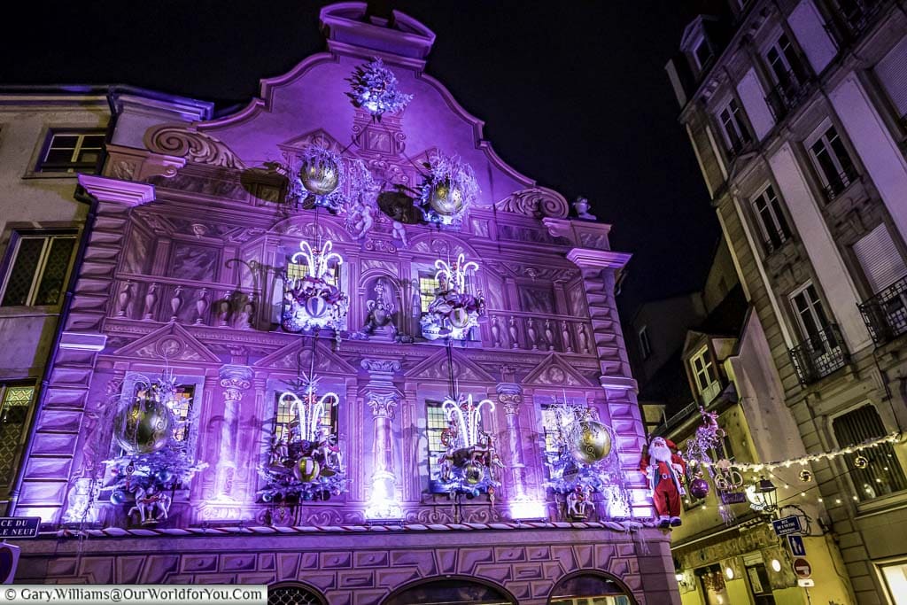 The ornately decorated gable end of a historic building brightly lit in pink with candy canes, reindeers, baubles and even Santa.