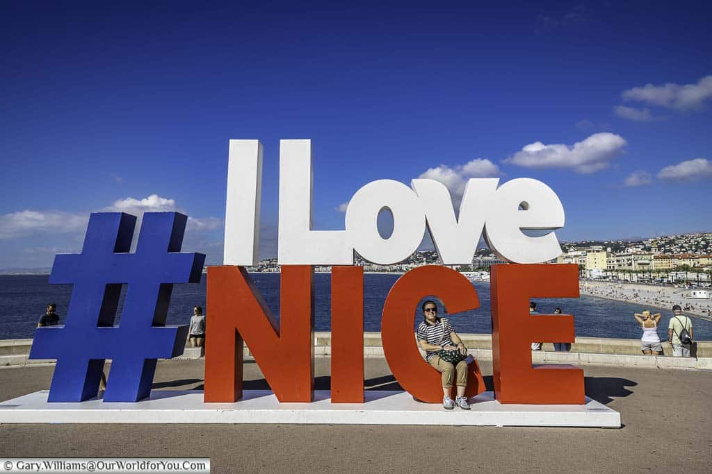 Janis seated on a giant hashtag I Love Nice sign overlooking the blue water of the Cote d'Azur in the south of France