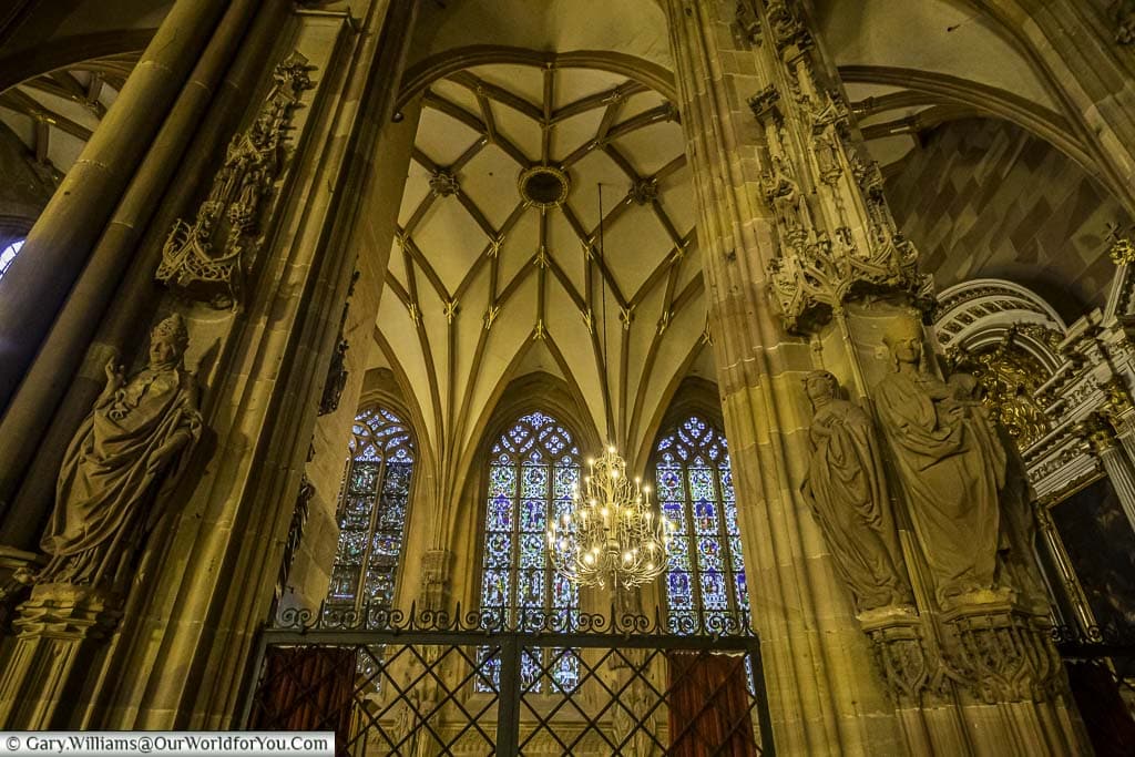 Inside the gothic Cathedral of Strasbourg looking through two pillars decorated with statues of the Virgin Mary and bishops towards a vaulted roof, from which an ornate chandelier is suspended in front of stained glass windows.