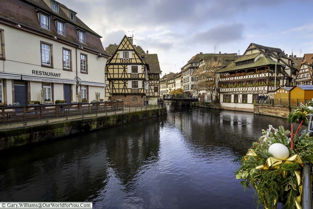 Overlooking the water in the Petite France region of Strasbourg with the waterside restaurant 'Maison des Tanners' with its decorated window boxes against the half-timbered building. You can tell it's Christmas at the handrails are decorated with baubles and wreaths.