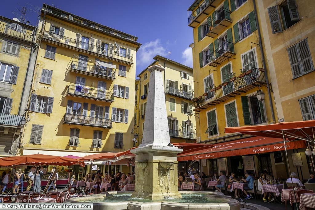 A fountain in a square in central Nice, surrounded by tables and chairs covered by parasols, Flanked on all sides by brightly coloured residential buildings.