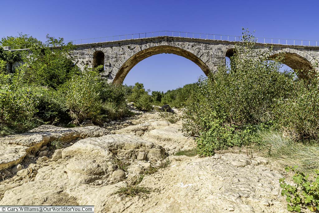 The small Roman bridge over a dry riverbed against a clear blue sky.