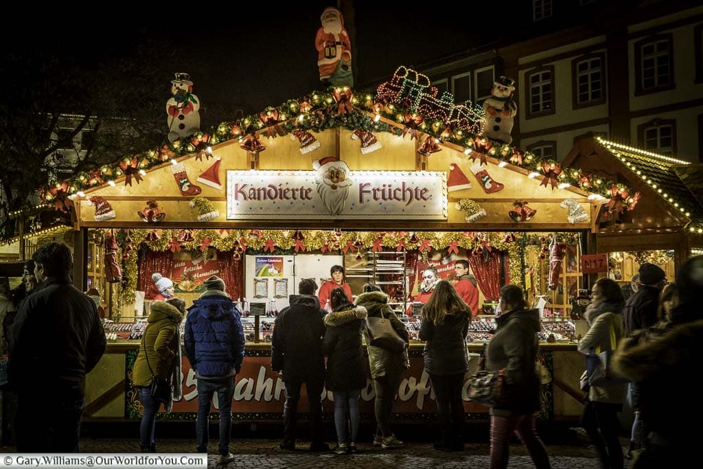 A brightly lit, candied fruit stall, on Frankfurt’s Christmas market. People gather to look at the selection of wares.