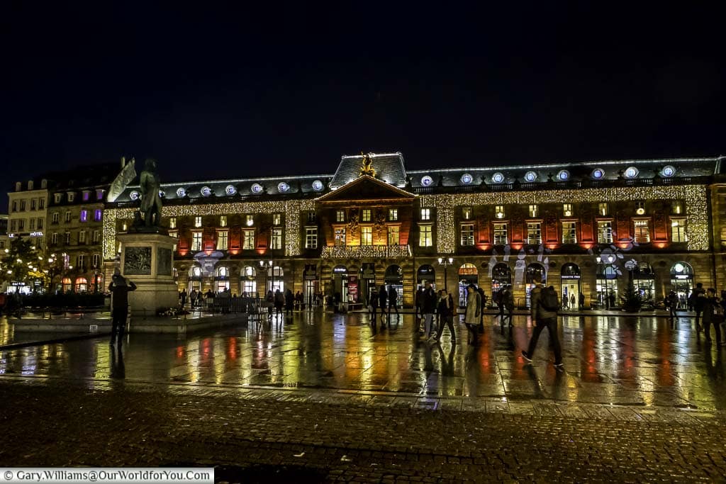 A scene across Place Kléber at night to the Aubette, a historic, mid-18th century, building brightly decorated in a mix of gold & red for Christmas. The square in front of the building, damp from some earlier rain, reflects the lights of the building.