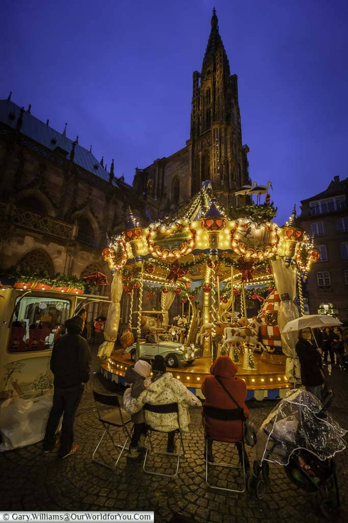 A family sitting around the carousel at the christmas markets in strasbourg at dusk in the shadow of the spire of its cathedral.