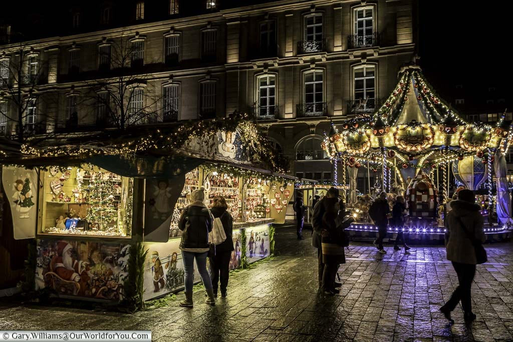 A small group of people gather between a Christmas market stall and the carousel in Place De La Cathédrale.