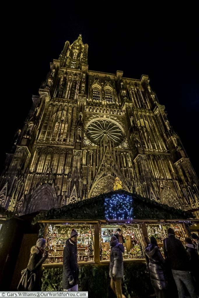 A low angle shot looking up at a Christmas market stall in front of the illuminated Cathedral of Strasbourg.