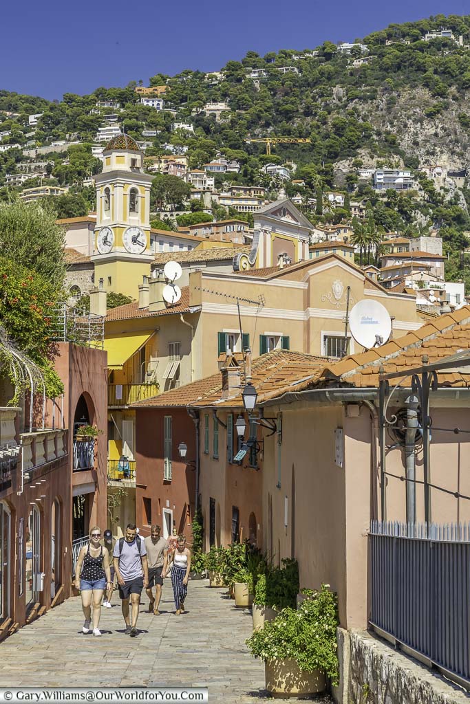 A pedestrian lane in Villefranche-sur-Mer lined with building painted in the bright colours, against the backdrop of the hills that surround the town.