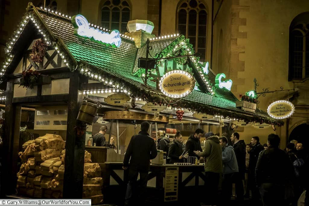 A food stall at night at the Christmas market in front of St. Catherine's Church. The food is cooked over an open wood-fired grill and you can see the sacks of logs stacked at the end of the cabin.