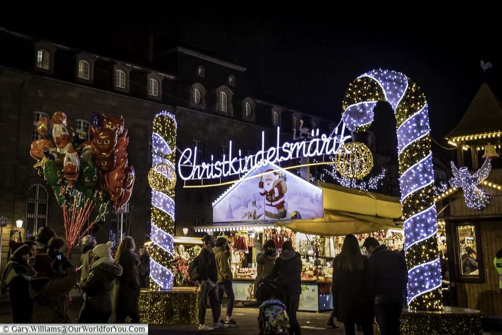 Folks mingling in front of the entrance to the Christkindelsmärik Christmas market, which is marked by a bright neon signed between two gold and silver striped candy canes created with fairly lights.