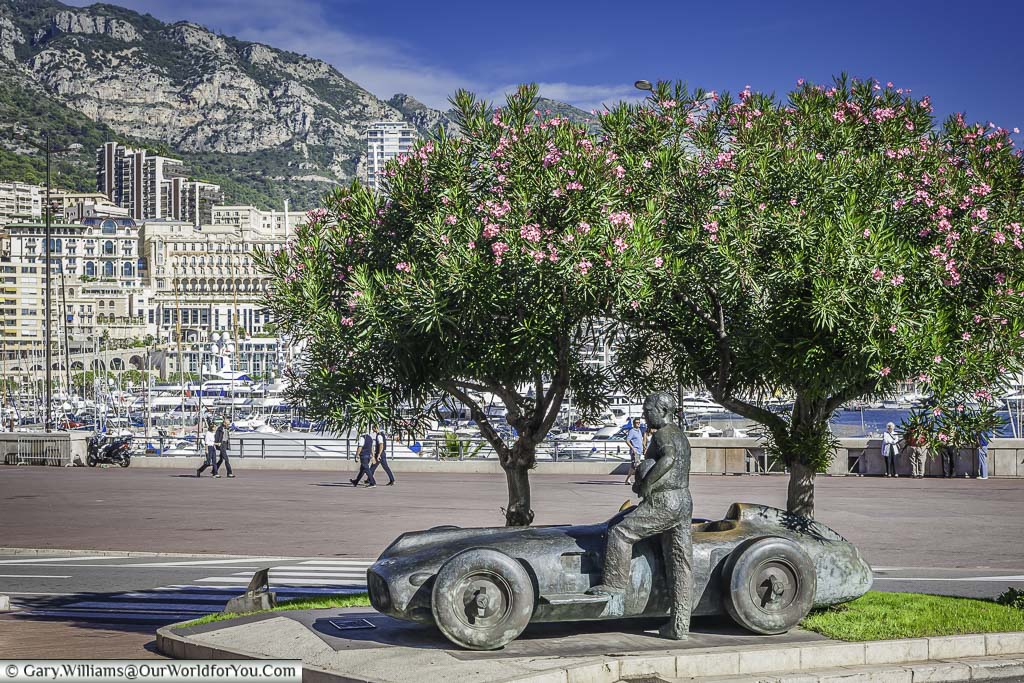 A bronze statue of a facing driver standing next to his 1950's formula 1 car under a couple of trees in flower, in front of the famous Monaco harbour.