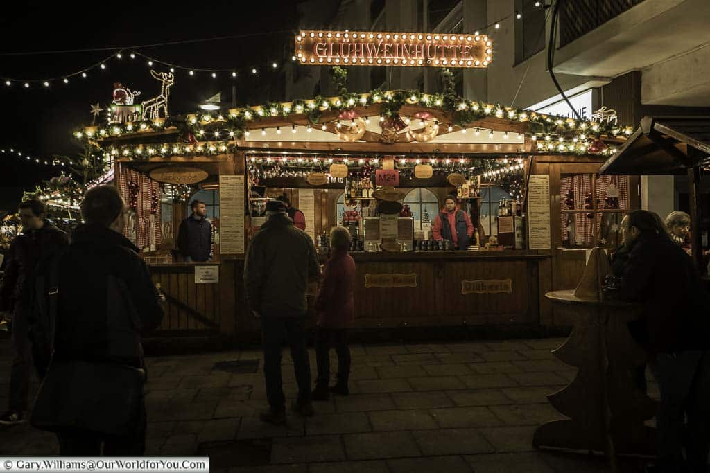 A brightly lit Glühwein hut in Frankfurt's quayside Christmas market at night. People are coming and going, others are perched around wooden tables, shaped like Christmas trees enjoying their drinks.