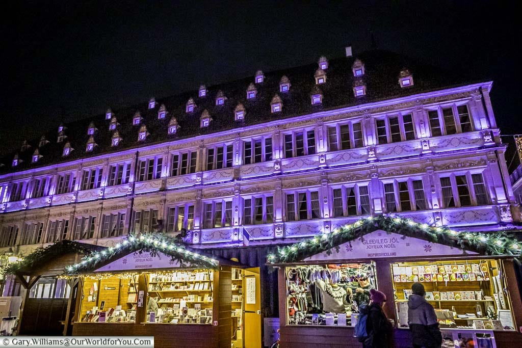 Christmas market stalls in the international market in Place Gutenberg in front of a historic building floodlit in a purple/lilac colour.