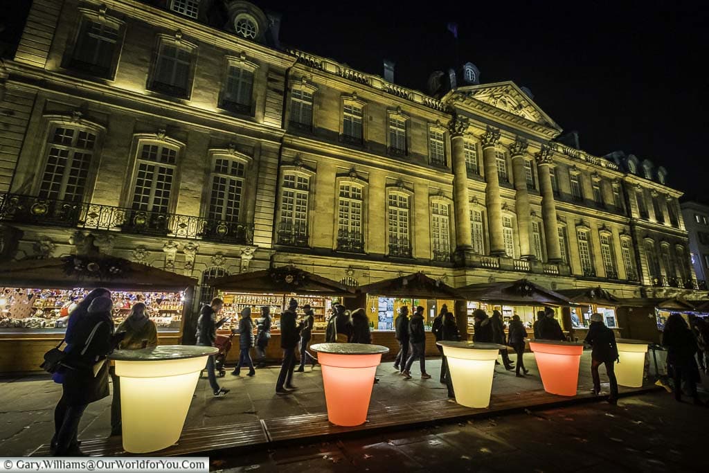 People gathering around illuminated tables that resemble giant plastic beakers in front of the Christmas market 'Marche-aux-Poissons' in front of the floodlit Palais Rohan.