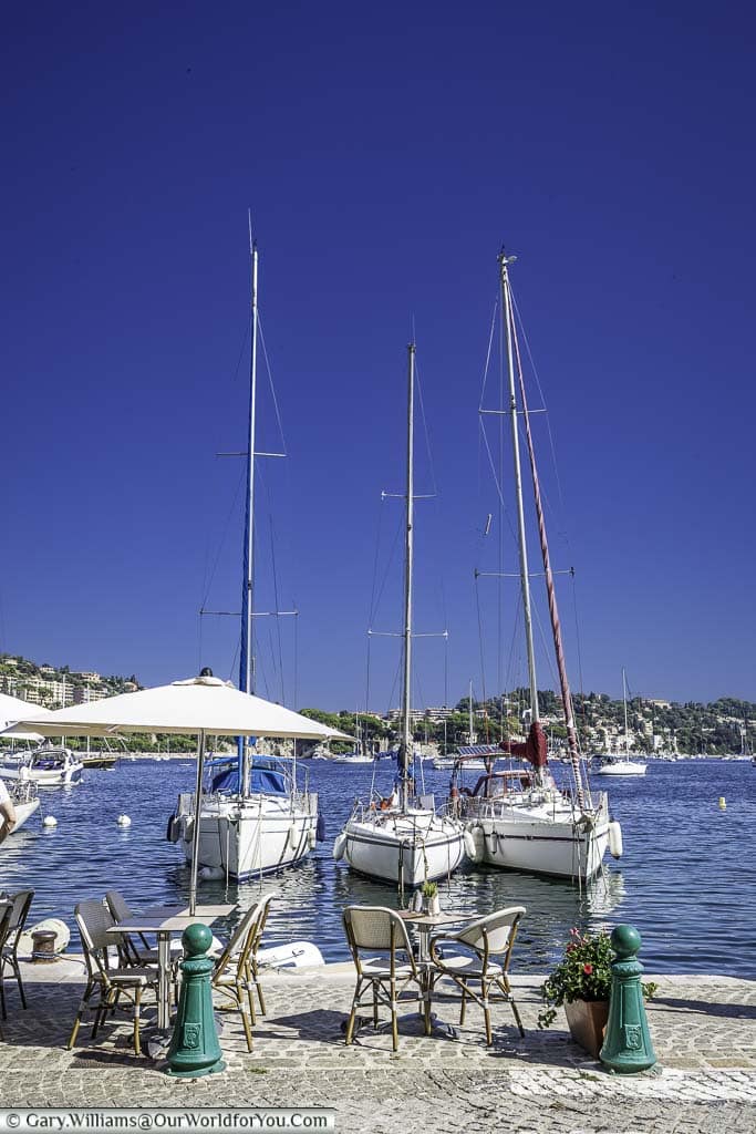 A table & chairs at the edge of the harbour of Villefranche-sur-Mer in front of 3 small yachts.