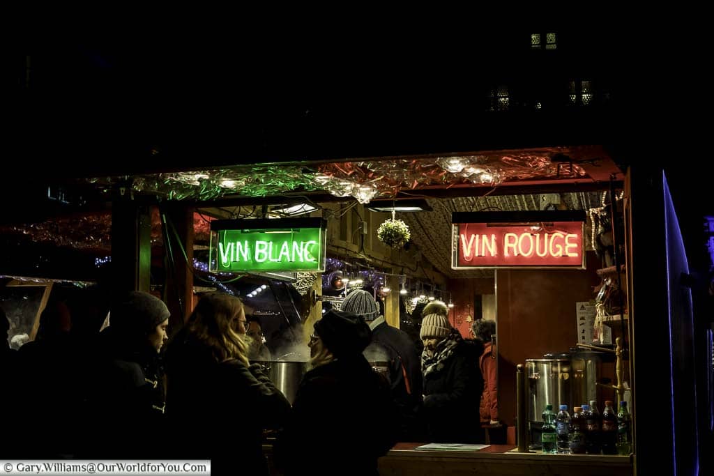 At the edge of a Christmas market drinks stall in the Place De La Cathédrale. There are two neon signs, one offering vin blanc, the other vin rouge.