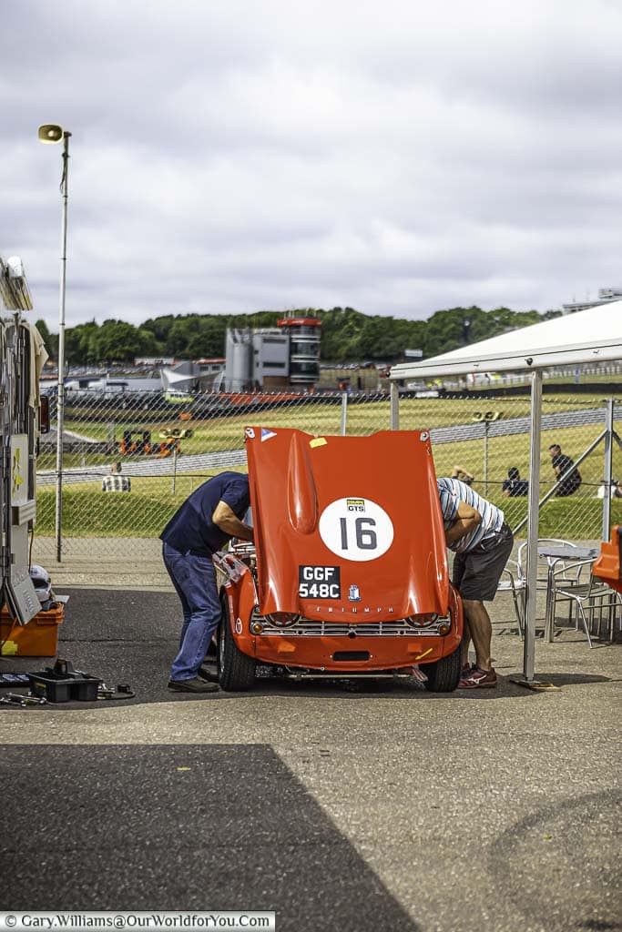 Two mechanics working under the raised bonnet of a classic, bright red, triumph tr4a in the paddock of brands hatch motor racing circuit in kent, england