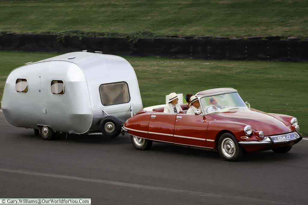 a bright red 1965 jcitroen ds 19 usine with its roof down towing a stainless steel caravan at the goodwood revival meeting