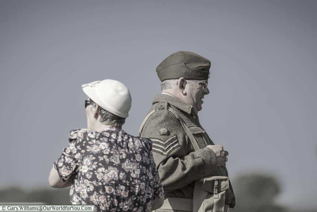 A couple dressed in period outfits, the man as a sergeant in the army, facing in opposite directions whilst looking on at the goodwood revival