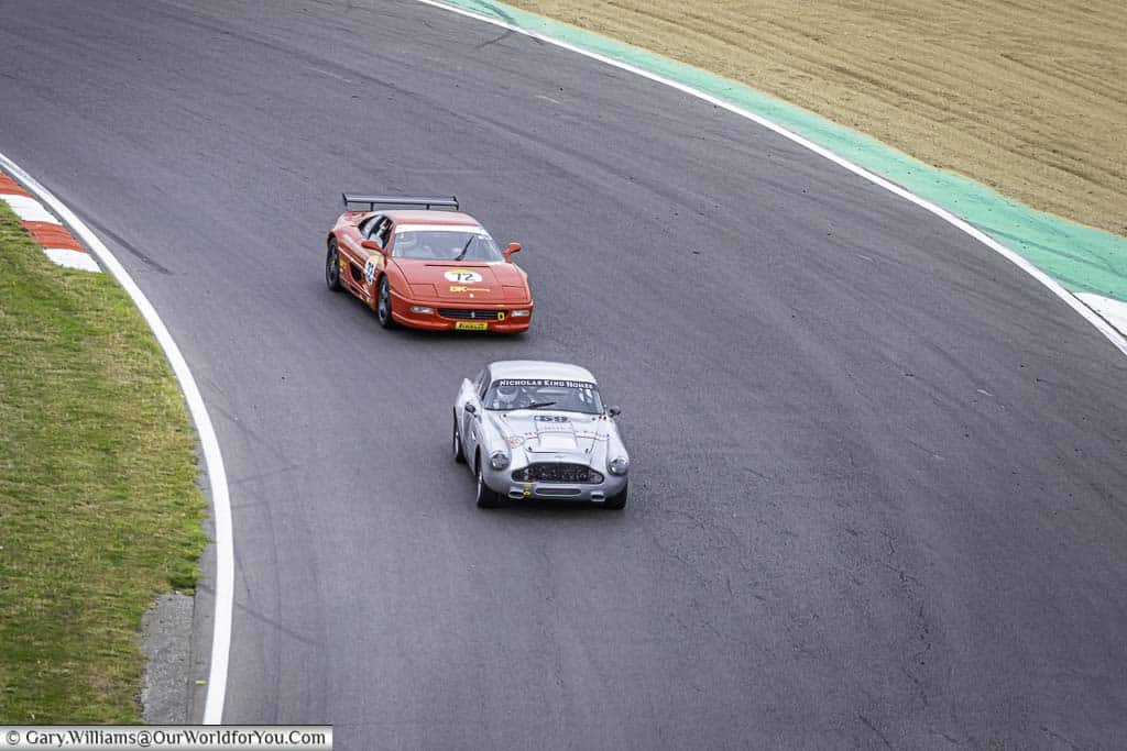 A red ferrari chasing down a classic aston martin as they drop down paddock hill bend towards hailwoods hill at brands hatch motor racing circuit in kent, england