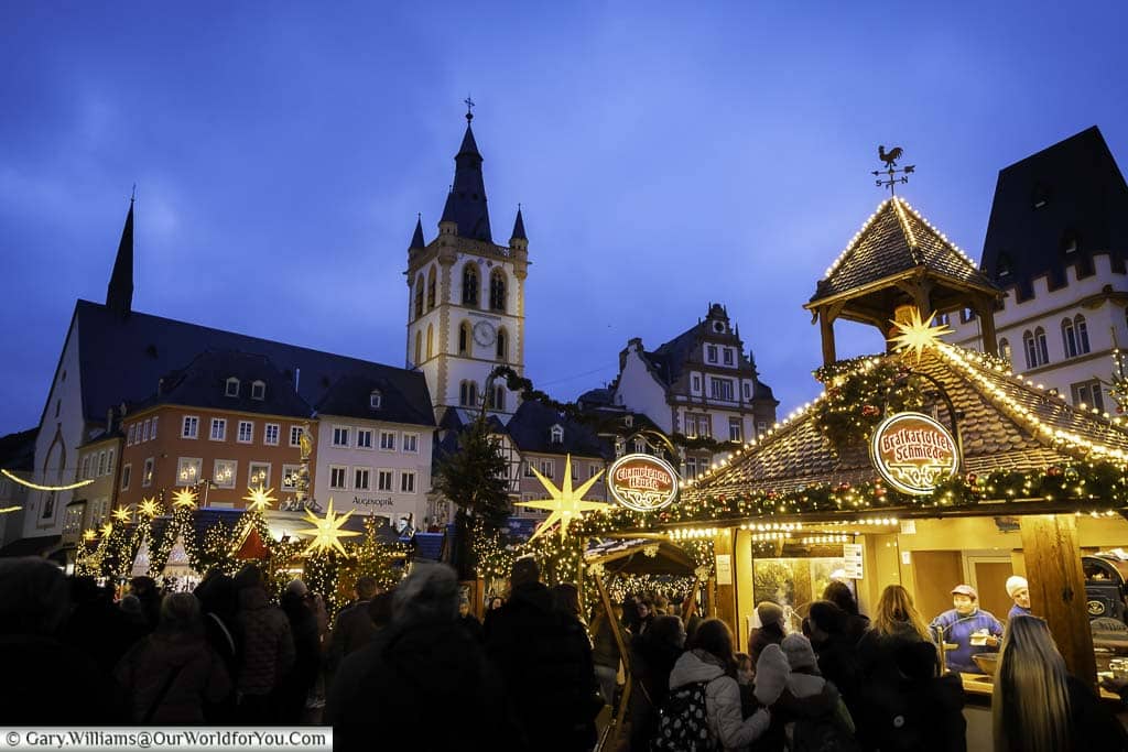 People gather at dusk around the edge of triers hauptmarkt for its Christmas markets lined with historic buildings and the tower of St Gandolf in the background.