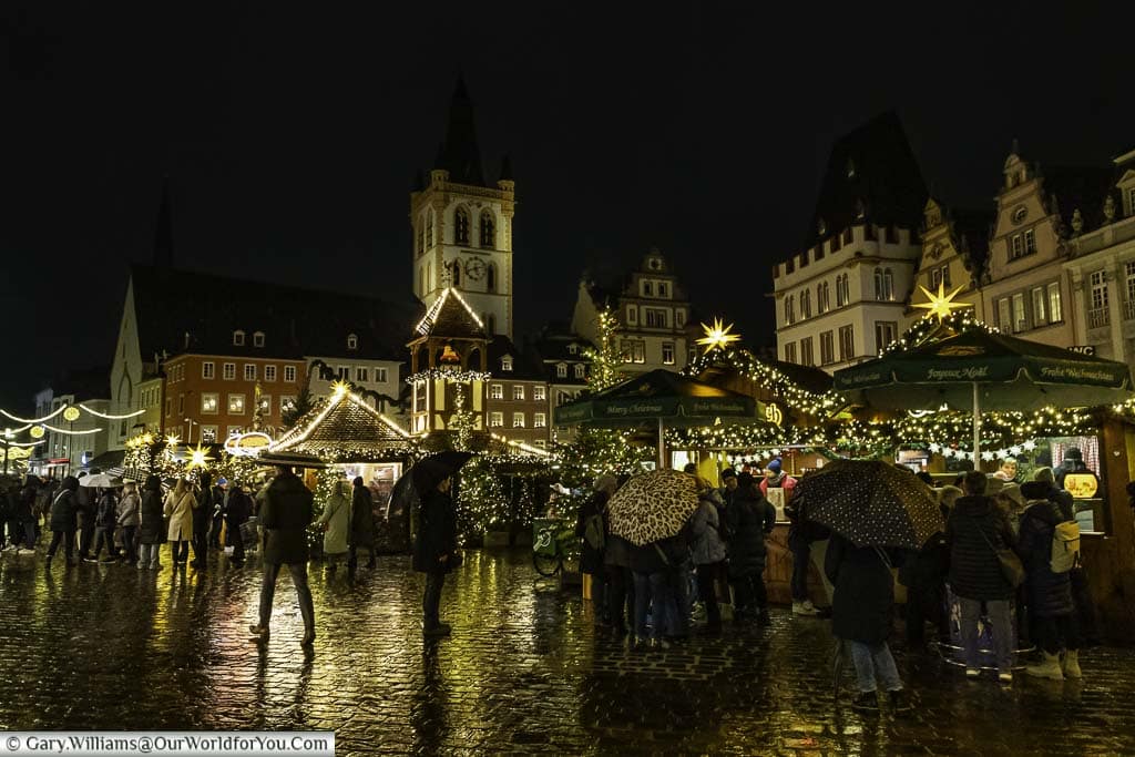 People, with umbrellas up, at the edge of triers hauptmarkt for its Christmas markets lined with historic buildings and the tower of St Gandolf in the background.