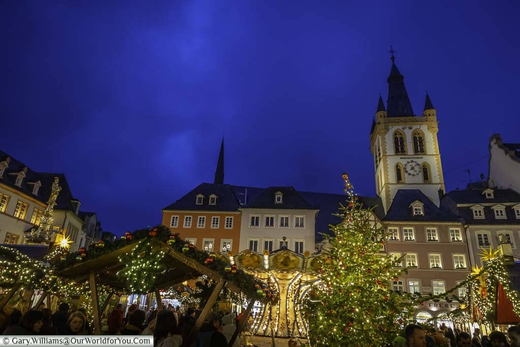 People gather at dusk in the centre of triers hauptmarkt for its Christmas markets lined with historic buildings and the tower of St Gandolf in the background.