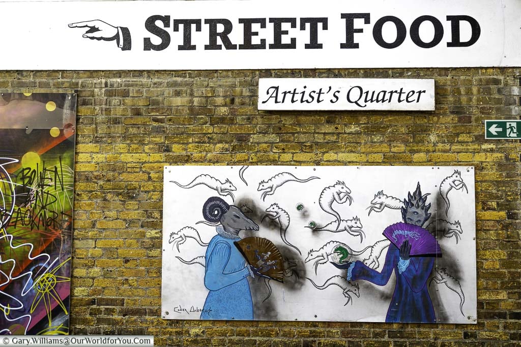 The wall on the artist's quarter in greenwich market featuring a murial and a sign pointing towards the street food section