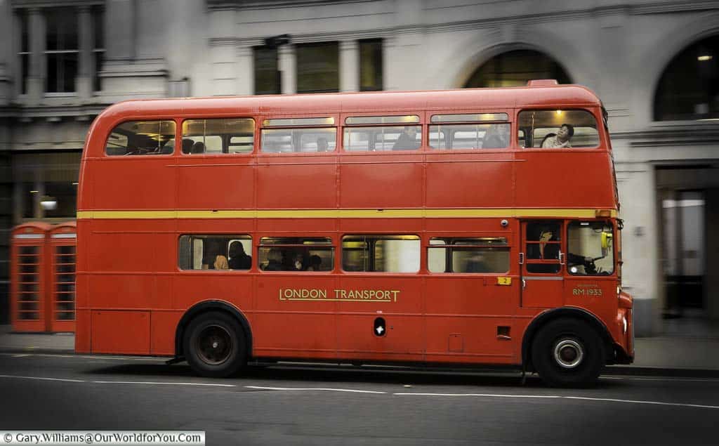 A selective toned image of a red London Routemaster bus passing 2 red telephone boxes against a Black and White street background.