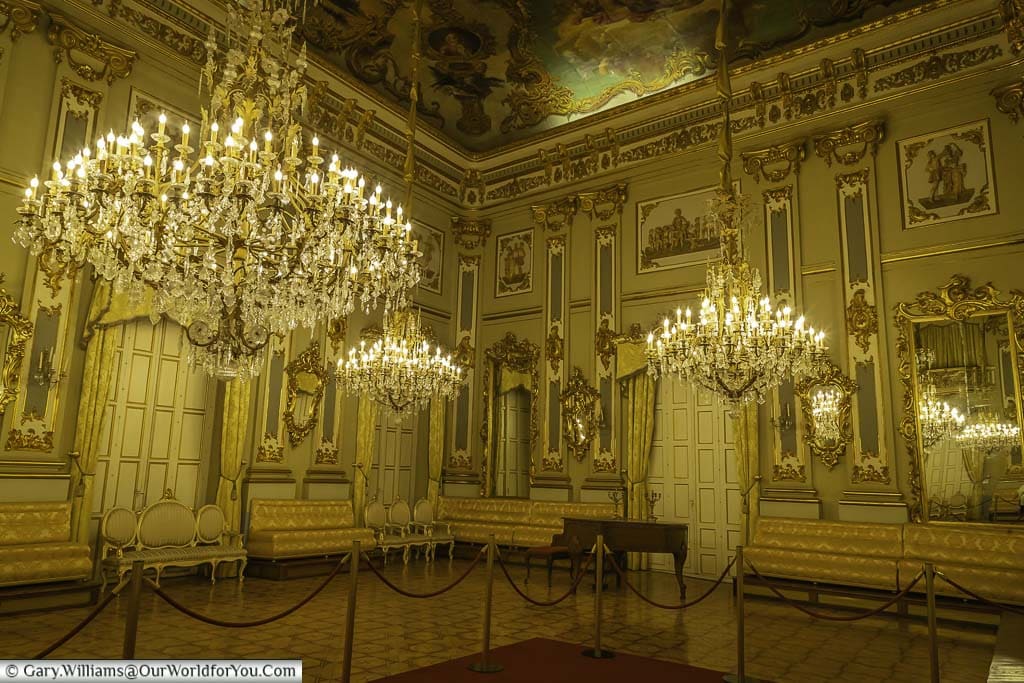 A photo of a grand ballroom in the Casino de Murcia with a red carpet on parquet flooring, flanked by rows of ornately decorated chairs. The ceiling is high and adorned with several large chandeliers.
