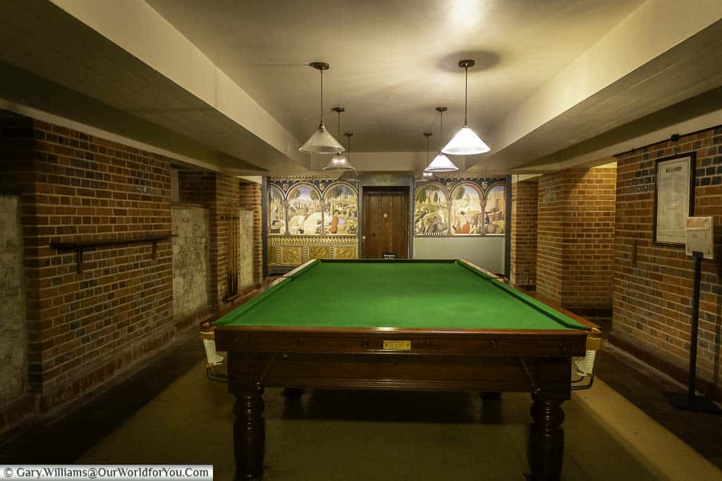 A brightly lit room with a green felt billiard table in the centre, illuminated by hanging light fixtures. The room has exposed brick walls and a colourful mural depicting medieval scenes on the wall behind the pool table.