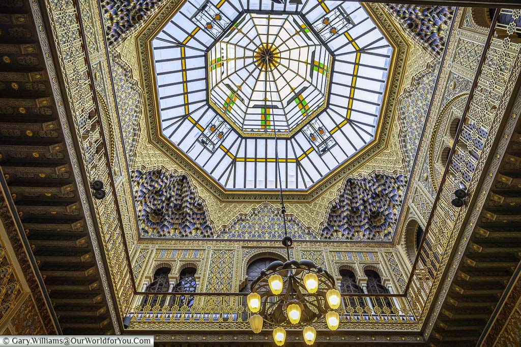 Looking up at a richly coloured stained glass dome ceiling in a mudejar design of the casino de murcia with an art deco chandelier in the centre. The dome is decorated with geometric shapes in green, blue, and yellow.